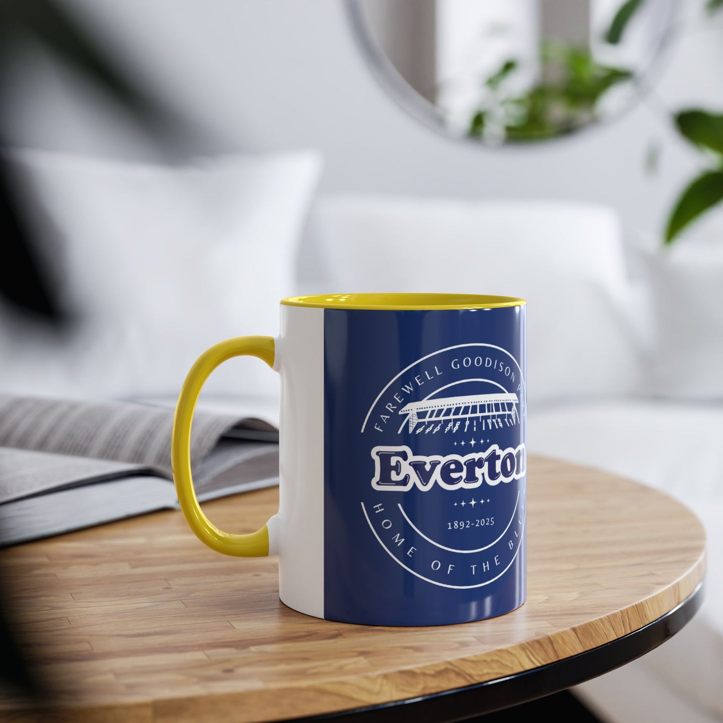 Lifestyle image showing the yellow infill mug in a living room environment it is on a wooden table with a blurred out book behind it and also a sofa with cushions, a mirror and a house plant are all blurred out around the mug image.
