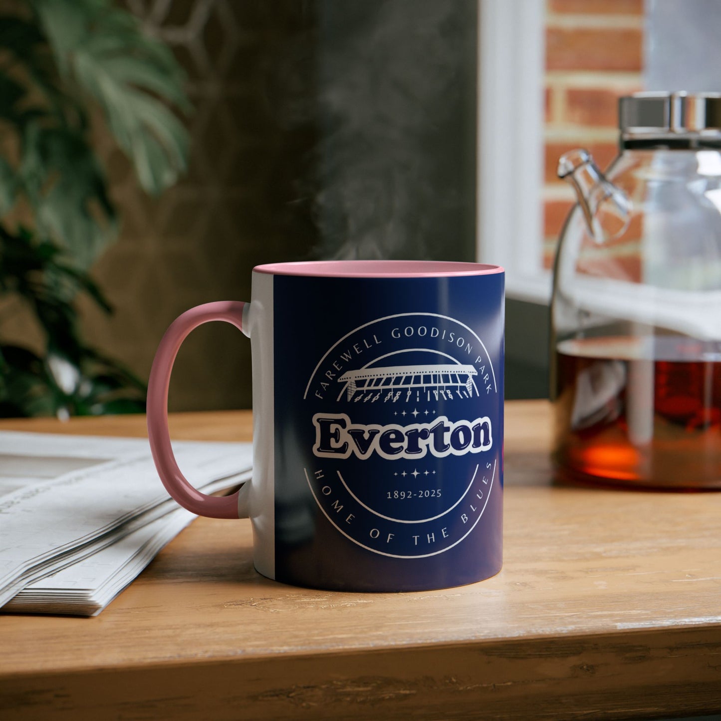 Lifestyle image shows the pink infill colour and handle on the left the main mug colour is navy blue with the graphic print on the front in white. The mug is placed on a wooden table with a newspaper next to it and a decanter of tea on the other side of the mug. This looks like a living room environment with a house plant and a window blurred out in the background.