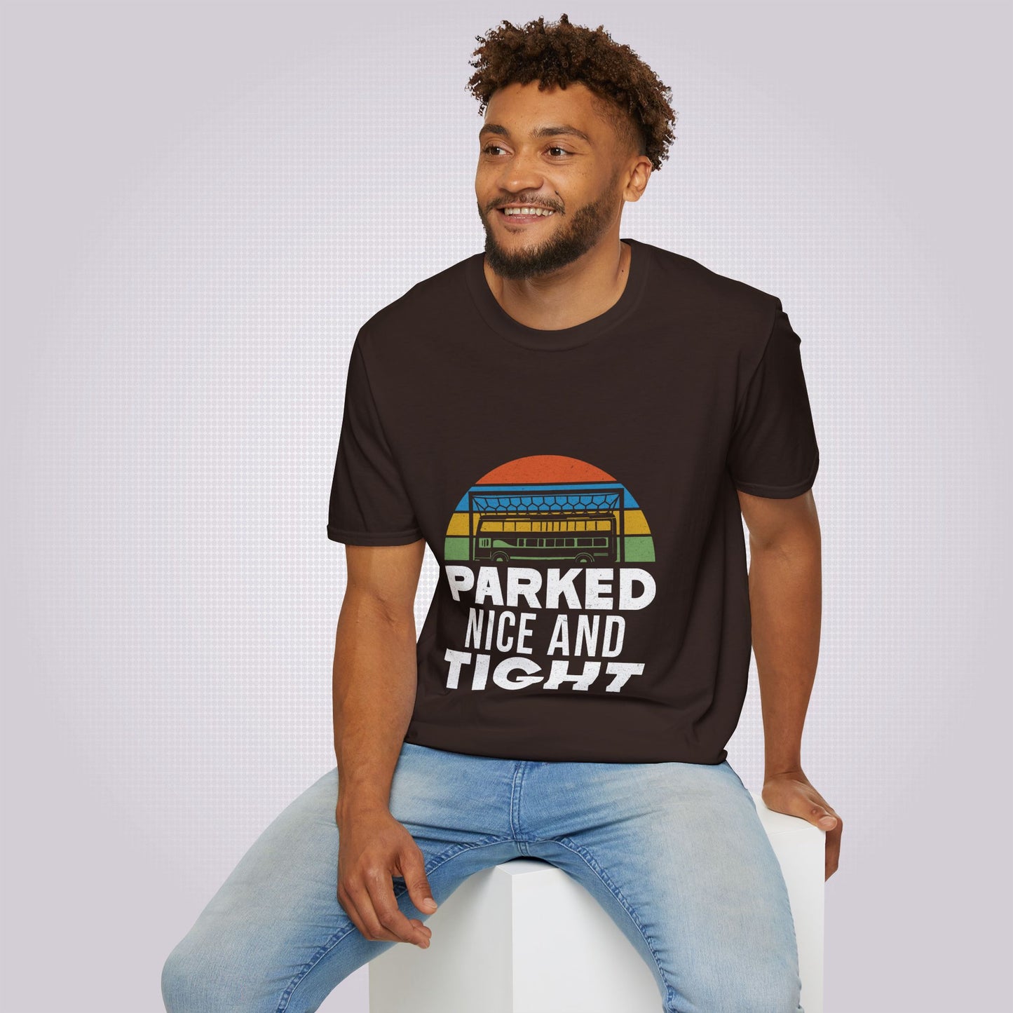Young mixed race male model sits on a white box wearing the brown t shirt with a pair of jeans he is looking at someone off camera and smiling at them