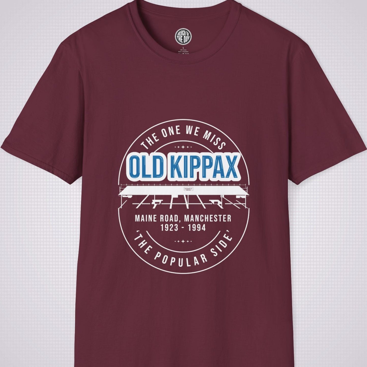 Maroon T Shirt on a light grey dotted background. There is an illustration of the Old Kippax stand as the focal point of the design, with the following text written above the drawing: 'The One We Miss' OLD KIPPAX. Then under the diagram it reads Maine Road, Manchester 1923 - 2004 followed by the words 'The Popular Side’