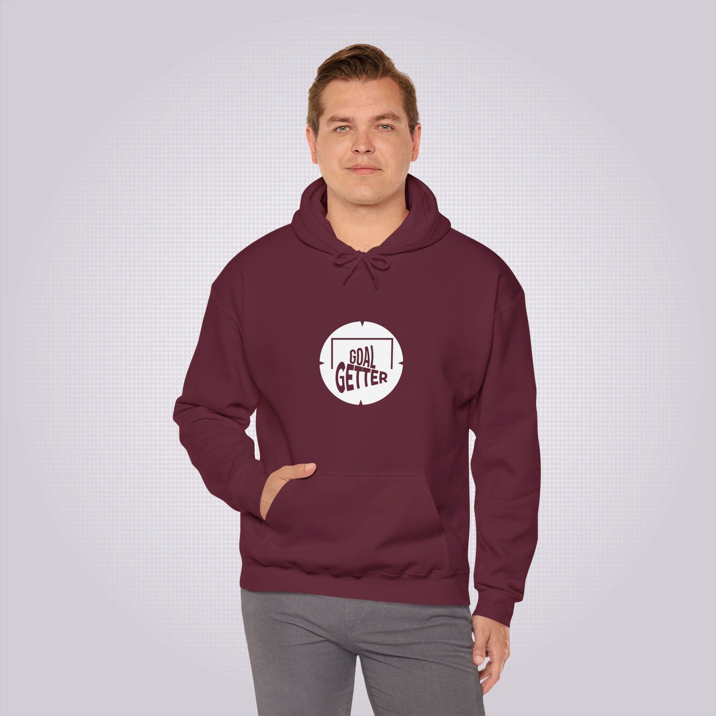 Young man with fair hair smiles at camera with one hand in the pouch pocket and the other by his side as he demonstrates the maroon hoodie with a pair of grey trousers