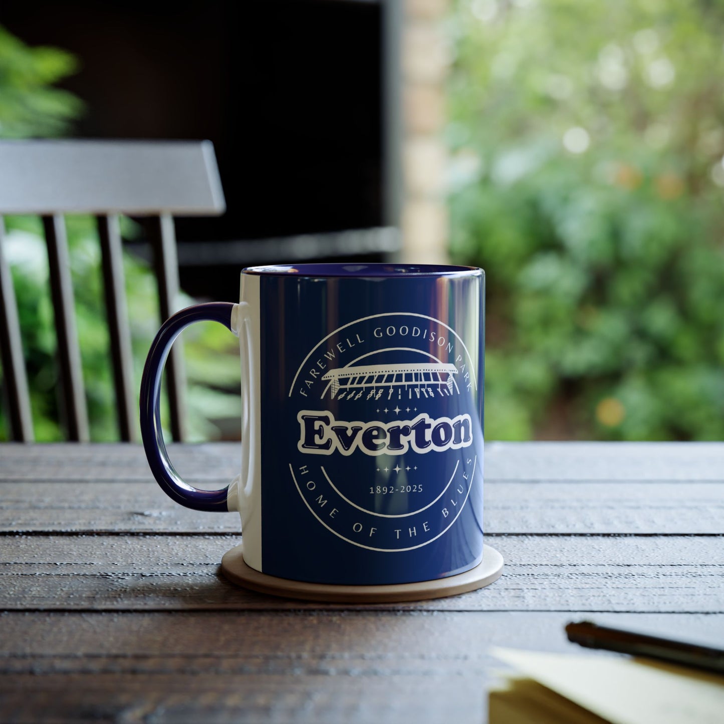 Lifestyle image shows the navy mug with the farewell goodison artwork on it, the mug is placed on a small wooden coaster on a wooden garden table, with a notepad and pen blurred out at the front, behind the mug there is a blurred out wooden garden chair and green foliage.