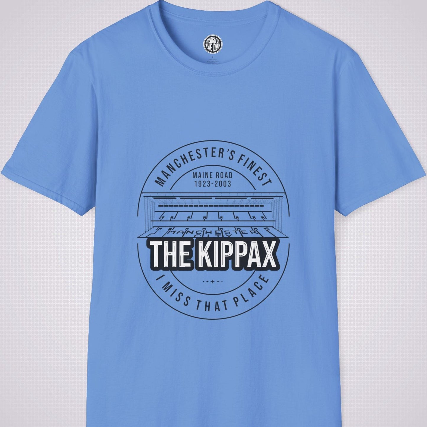 Mid Blue T Shirt on a light grey dotted background. There is an illustration of the Kippax Stand, above the illustration it reads 'Manchester's Finest Maine Road 1923 to 2003'. Below the illustration it reads 'The Kippax I miss that place'