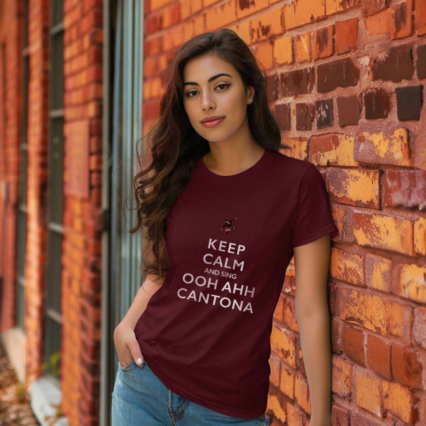 Lifestyle image of an attractive young woman wearing the burgundy t shirt with a pair of jeans she is stood against a wall with a hand in her pocket