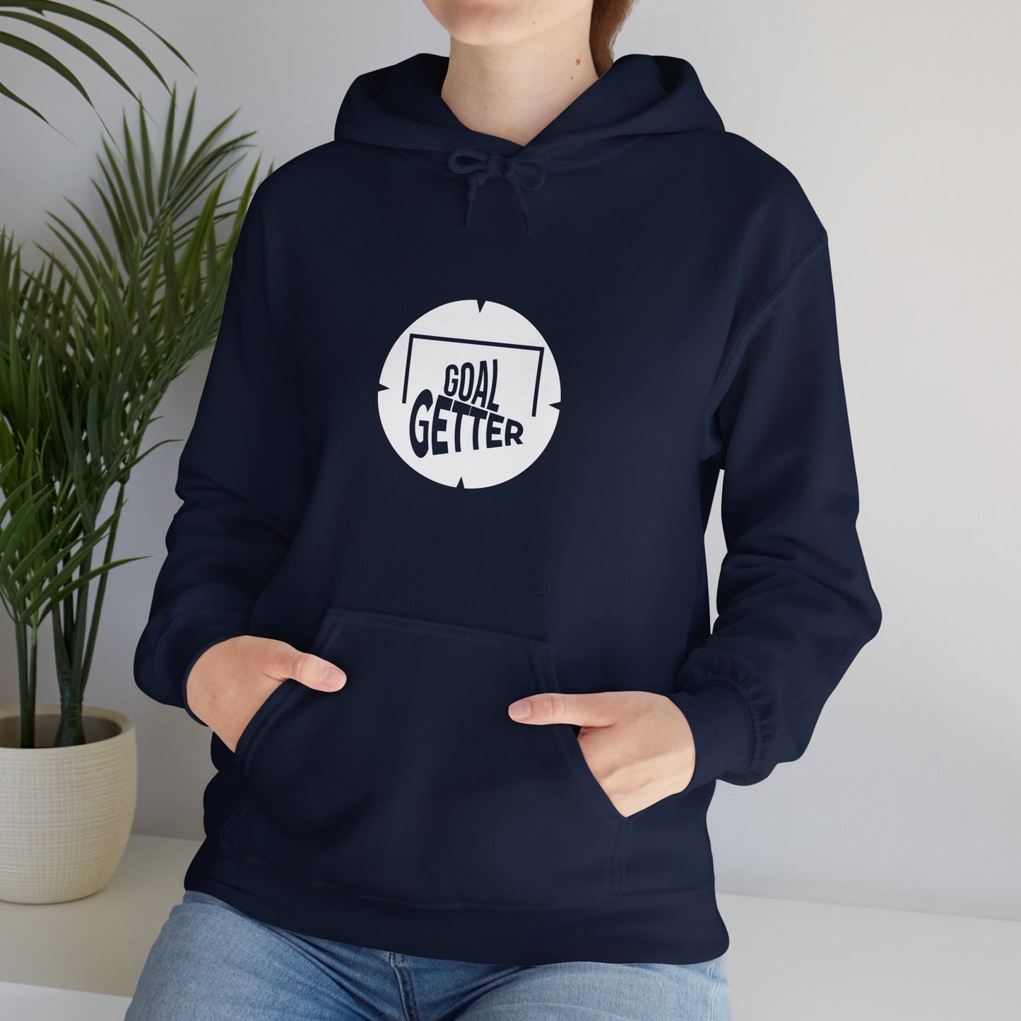 Lifestyle shot of a young woman sitting on a white shelf with a house plant next to her she is wearing the navy hoodie with jeans