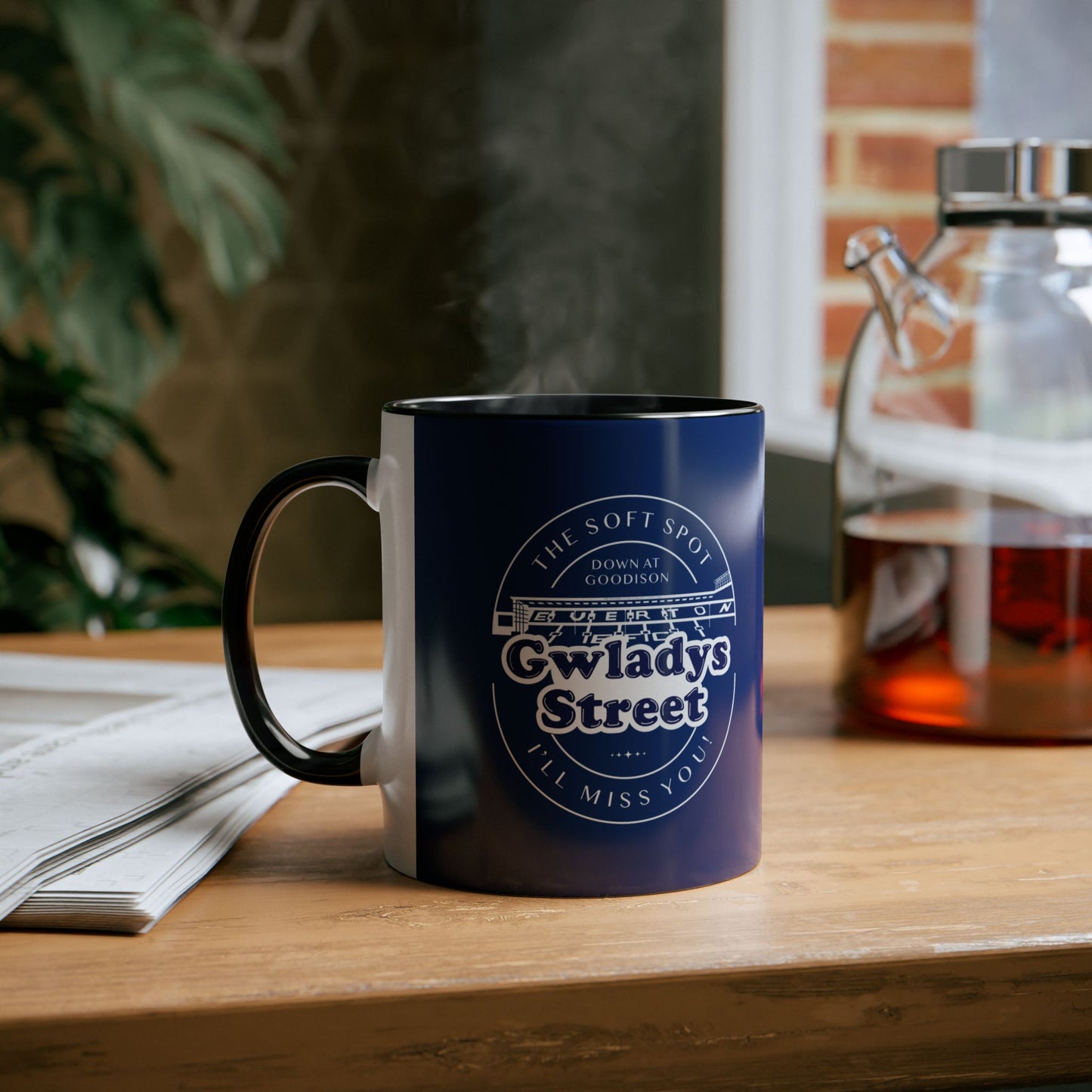 Lifestyle image which shows the navy blue mug with black handle and inners on a wooden bench with a flask of tea and a newspaper and houseplants blurred out around it