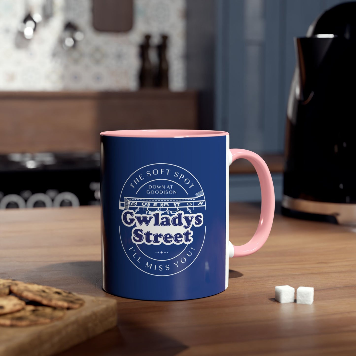 Lifestyle image shows the navy blue and pink mug in a kitchen environment. The mug is placed on a laminate wood worksurface, it has a kettle, some sugar lumps, a chopping board with cookies on it, and a cooker all blurred out around the mug image.