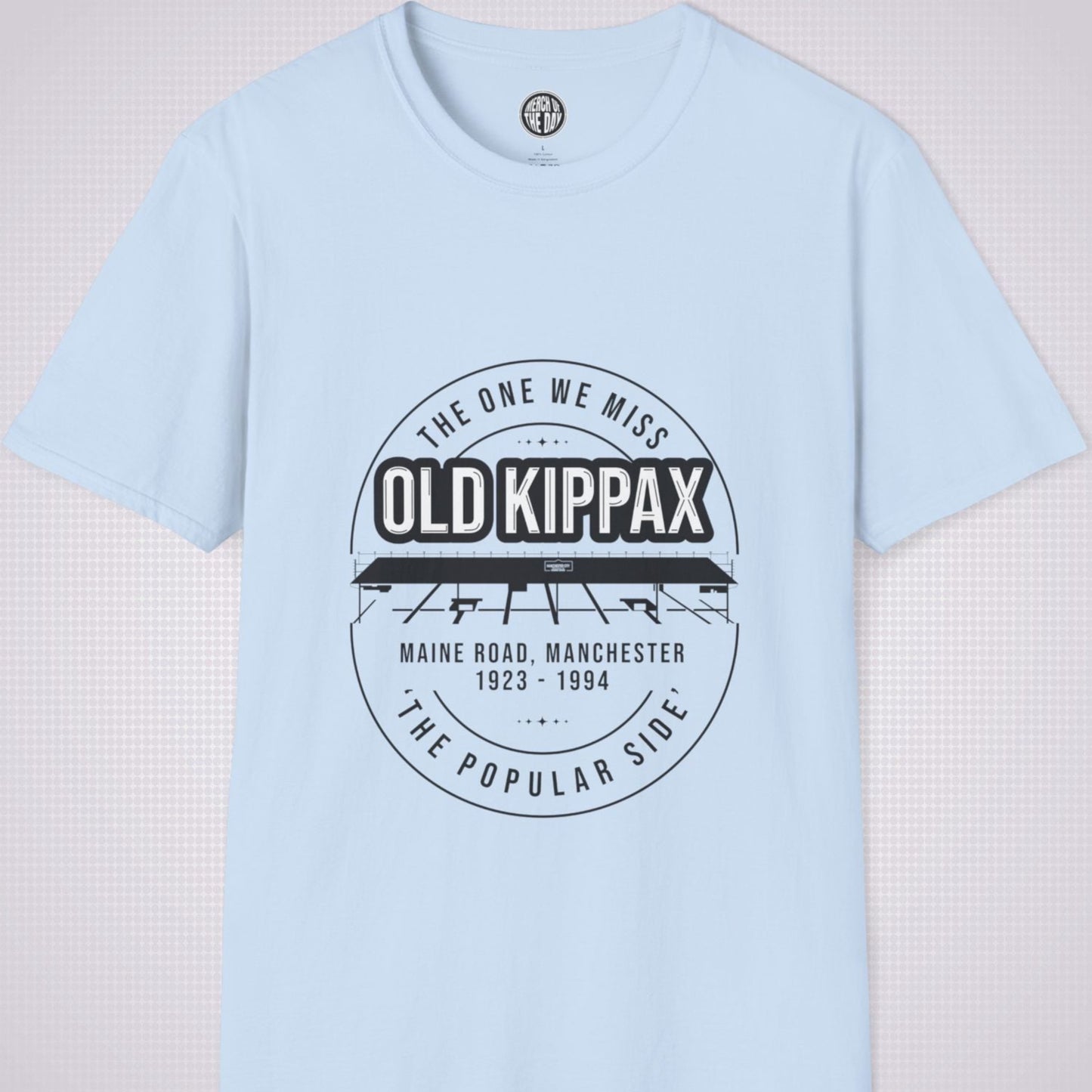 Light Blue T Shirt on a light grey dotted background. There is an illustration of the Old Kippax stand as the focal point of the design, with the following text written above the drawing: 'The One We Miss' OLD KIPPAX. Then under the diagram it reads Maine Road, Manchester 1923 - 2004 followed by the words 'The Popular Side’