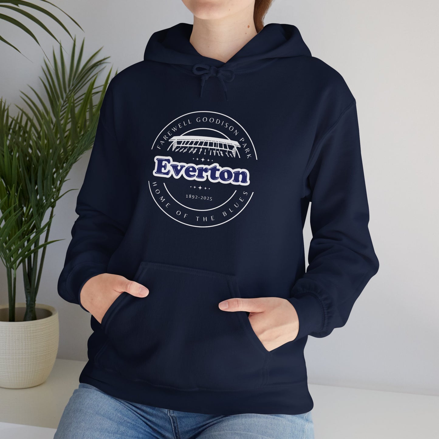 Lifestyle image showing a woman wearing the navy hoodie with a pair of jeans she is sat on a white shelf with a house plant next to her she has her hands in the pouch pocket demonstrating the design of that feature