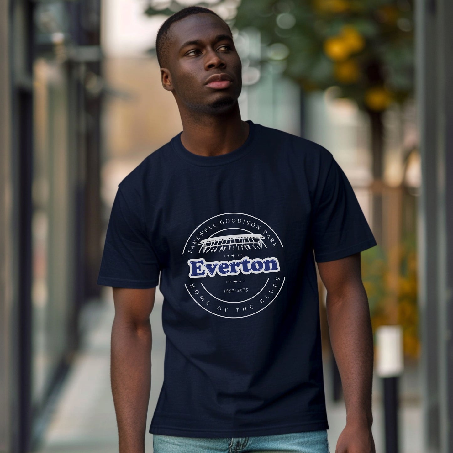 Lifestyle image of an African male walking down what looks like a city street there are blurred buildings and trees in the background he is looking to the left as you look with his body facing the camera and he is wearing the navy t shirt