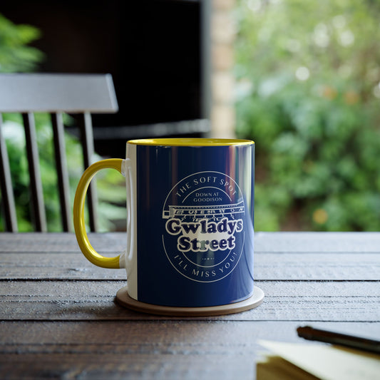 Lifestyle image of the Navy blue and Yellow mug in an exterior setting with a wooden table and garden chair, a pen and notebook and dark green foliage all blurred out around the mug image