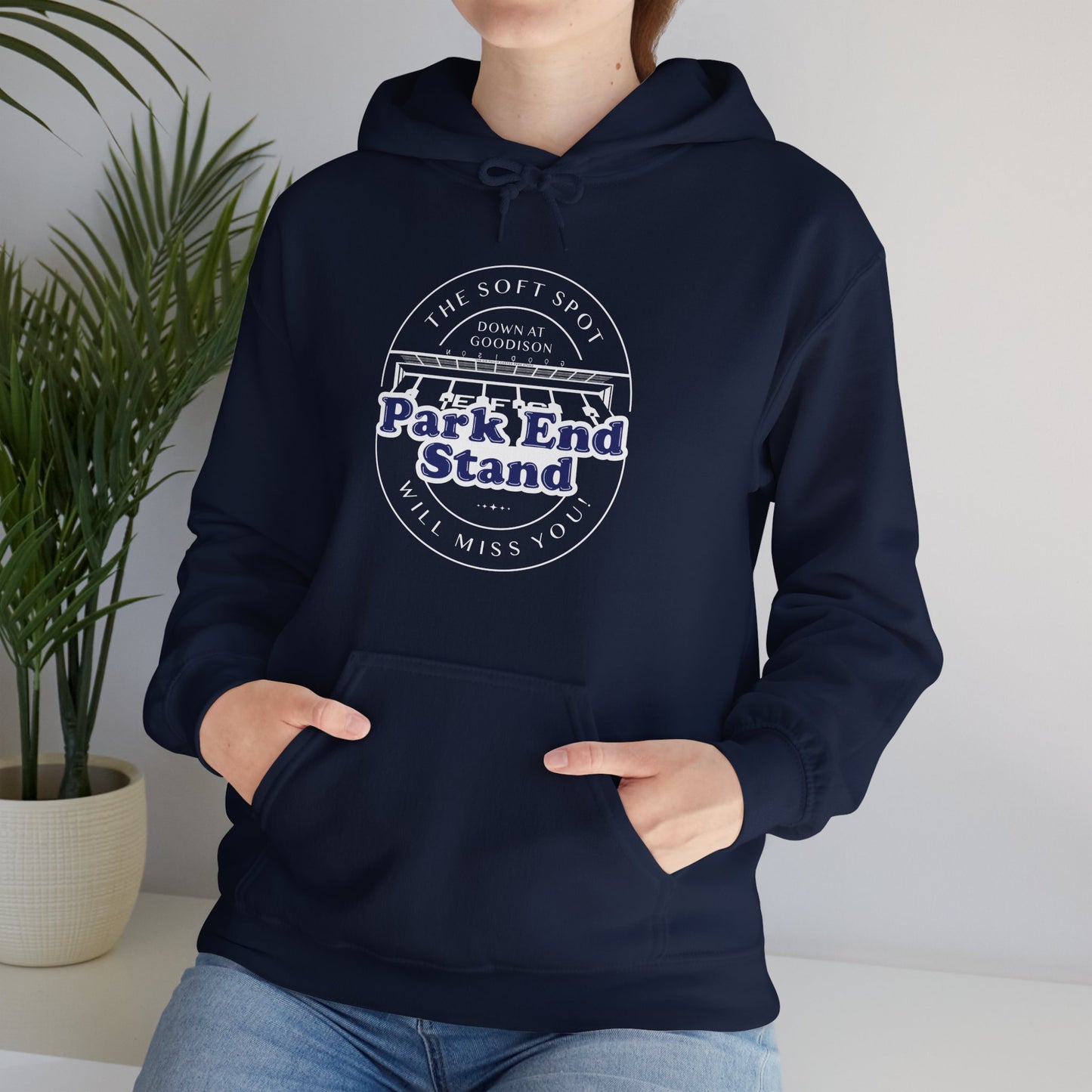 Lifestyle image shows a woman sitting on a white shelf with a house plant next to her she is wearing the navy hoodie with a pair of jeans and her hands are in the pouch pocket