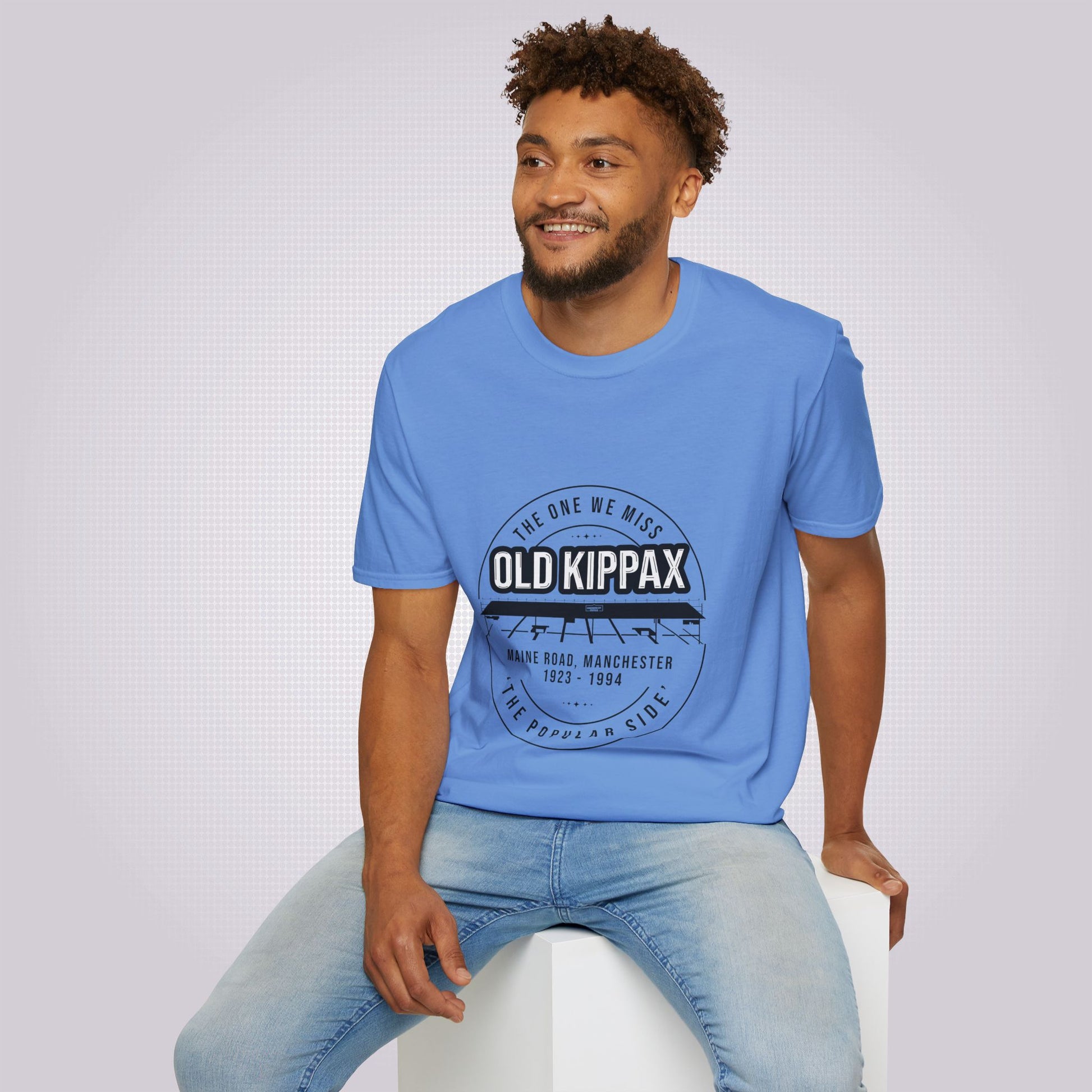 Young mixed race man in his twenties models the mid blue t shirt with a pair of jeans as he sits on a white box looking at someone off camera and smiling.