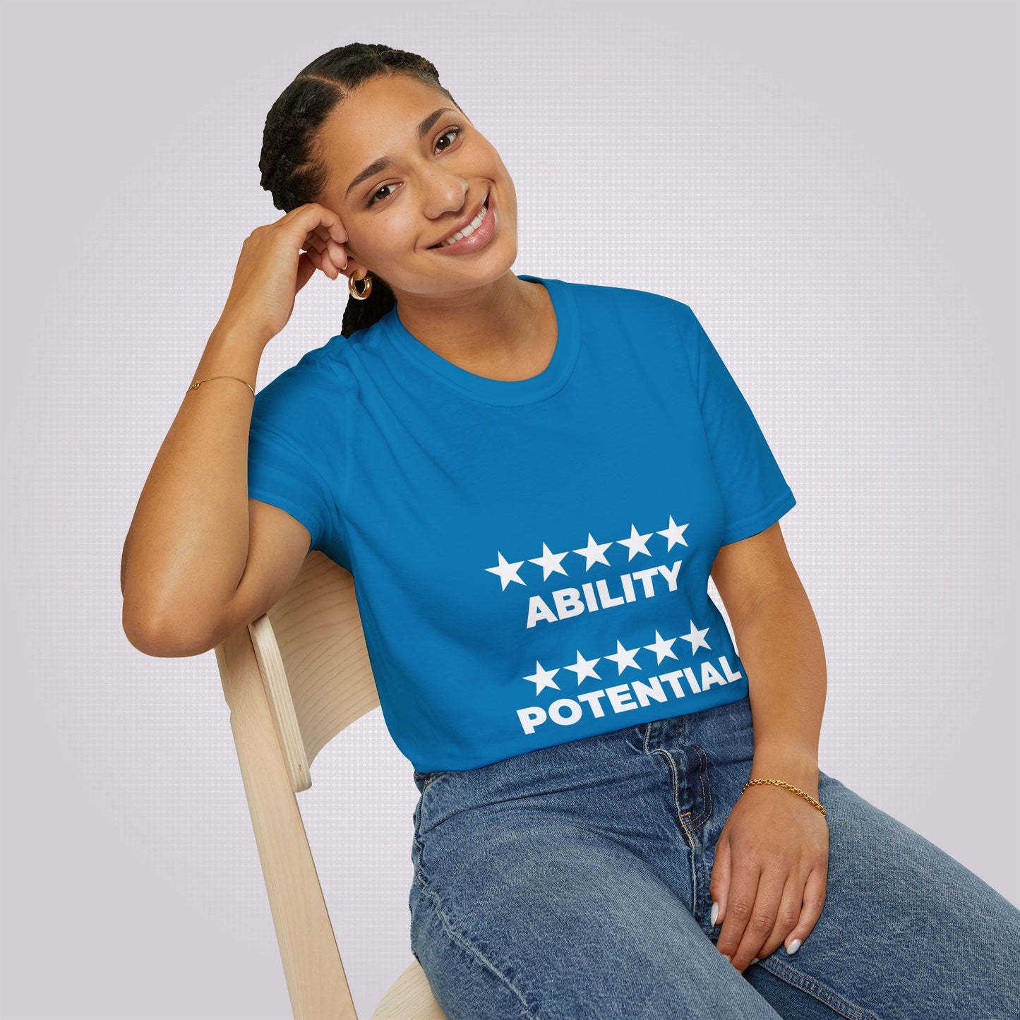 Attractive young mixed race woman in her twenties sits on a chair wearing the royal blue t shirt and jeans she is leaning on her elbow and smiling at the camera