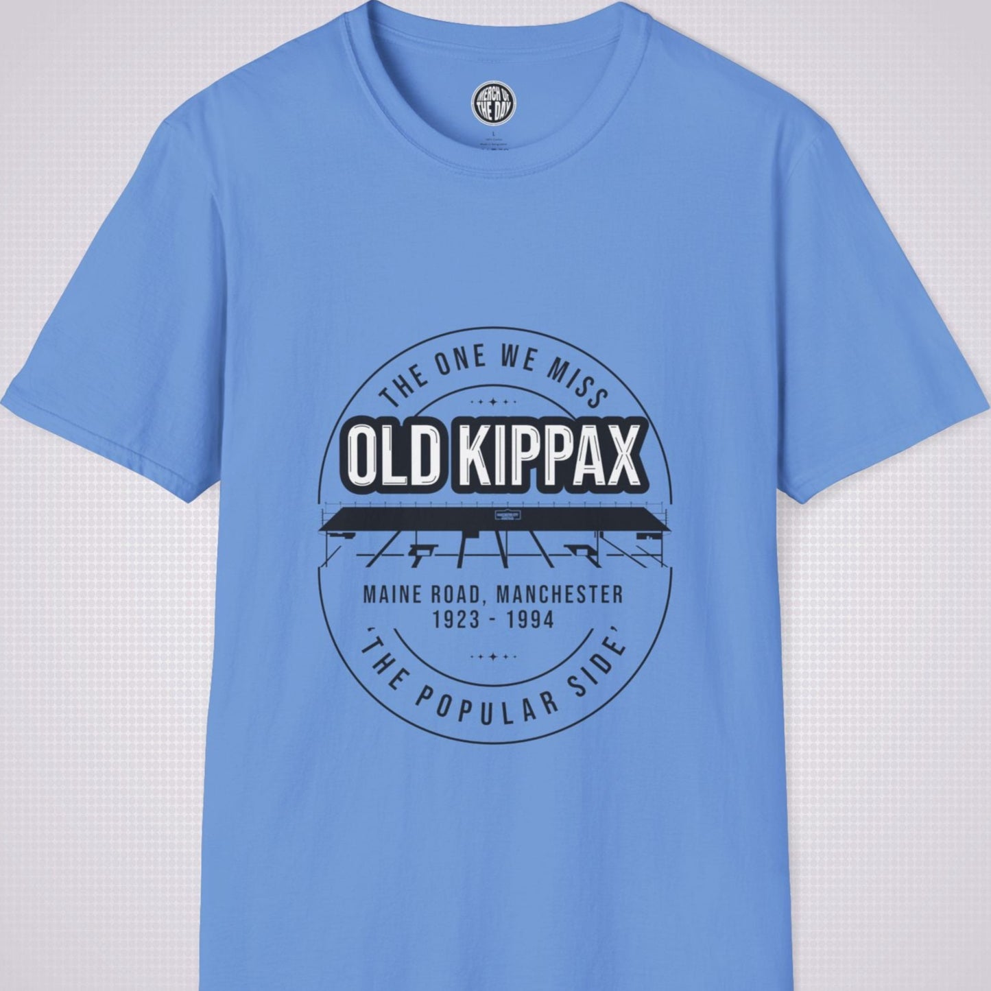 Mid Blue T Shirt on a light grey dotted background. There is an illustration of the Old Kippax stand as the focal point of the design, with the following text written above the drawing: 'The One We Miss' OLD KIPPAX. Then under the diagram it reads Maine Road, Manchester 1923 - 2004 followed by the words 'The Popular Side’